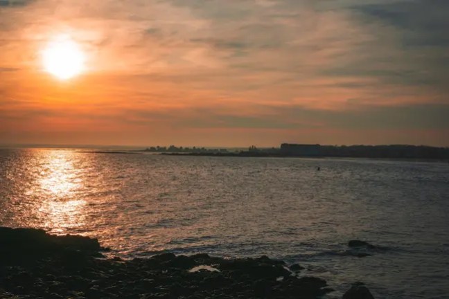 Sunset over ocean with rocky shore and distant horizon.